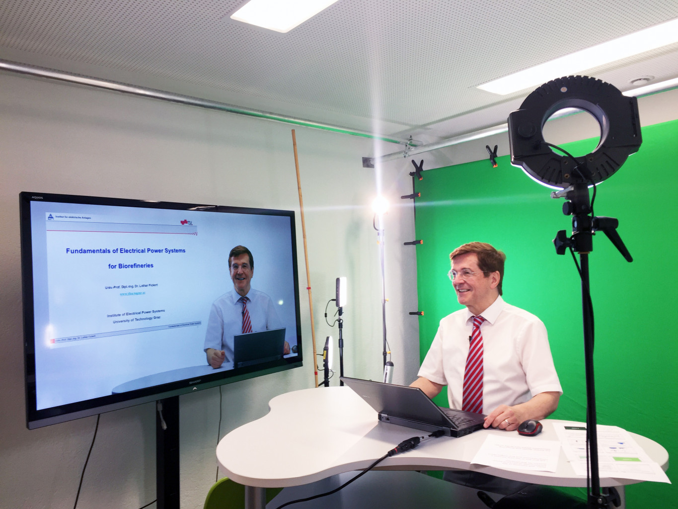 A person standing at a desk in the video studio, behind them is a green screen. Next to them is a preview of their slides.