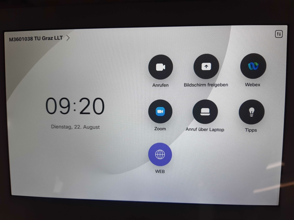 photo of the touch panel. You can see the name of the system, the time and date, and the menu items Call, Share, Webex, Zoom, Call via Laptop, Tips, WEB. The interface is in German.