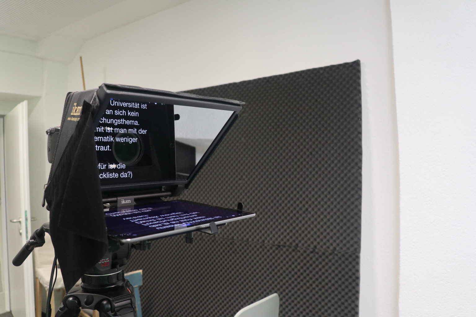 Teleprompter on a tripod in the studio, the script running on the mirror screen, an acoustic panel on the wall behind it